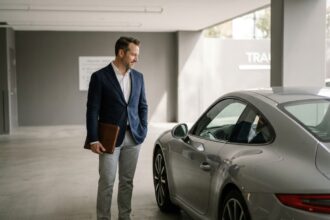 Homme en blazer marine consultant des documents d'assurance devant une Porsche 911 argentée dans un garage premium